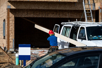 New home construction site, workman in red hardhat carrying length of wood board, housing development project
