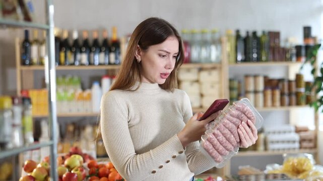 Girl scan QR-code on raw pork sausages package in store. Market visitor choose consider raw pork sausages semi finished products. Buyer pick up meat ware for bbq