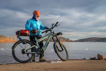 Obraz premium Senior cyclist with a touring e-bike with action camera mounted on handlebar at shore of frozen Horsetooth Reservoir in Colorado foothills.