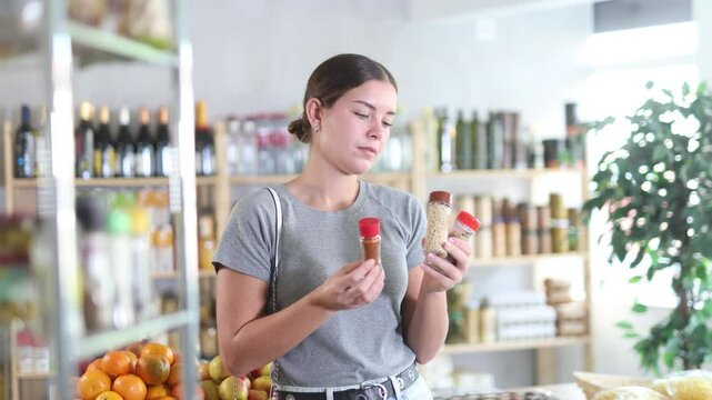 In healthy eating store, young woman buyer choosing spices items. Clients look package of spices bottle, inspect product and examines contents of package, compare paprika and chopped red pepper. 
