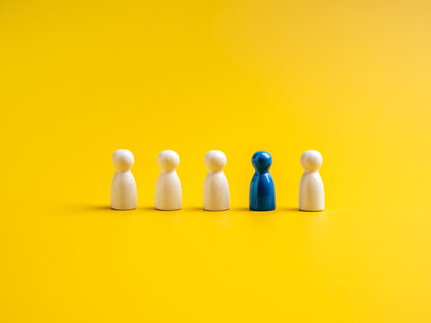 The blue one, different standing out of the group of white peg dolls, people symbols, on yellow background. Stand out from the crowd, leadership, unique, influencer, talent people, difference concept.