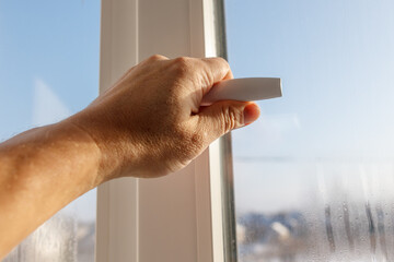 A hand is holding a white chalk stick in front of a window