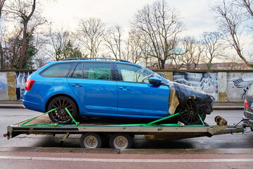 Damaged blue car on tow truck for transport at city street. Crashed vehicle covered in plastic film...