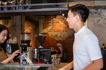 Fototapeta premium Asian female barista in apron assists smiling male customer at café counter with modern interior and exposed brick wall, showcasing friendly service and digital ordering interaction