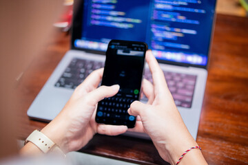 Close-up of Asian hands using smartphone while laptop screen displays coding interface on wooden café table, showing multitasking between messaging and programming in a modern coffee shop setting.