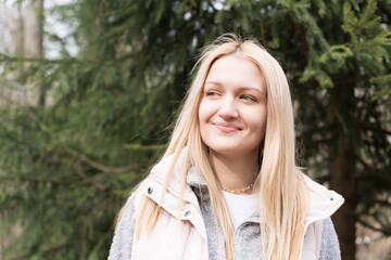 A blonde woman with long hair in casual clothes stands in a lush green forest, looking into the...