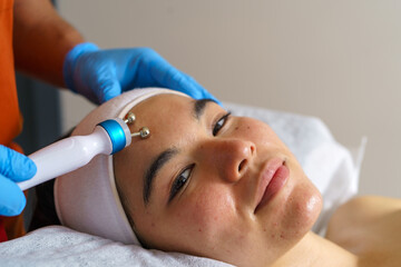 Beautician using a modern electric device for an acne treatment on the forehead of a young Asian woman lying on a bed