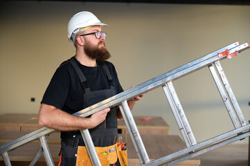Construction worker holding ladder. Man carrying steel ladder. Safe at work. Bearded builder standing in warehouse wearing hardhat. Repairs concept. Construction builder worker carrying ladder © Василь Івасюк