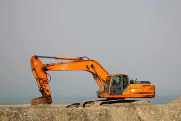 An orange excavator stands on a construction site by the sea. Its boom is outstretched and the...