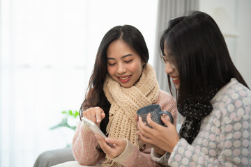 Two young women enjoying a cozy moment together while browsing on a smartphone, sipping hot drinks,...