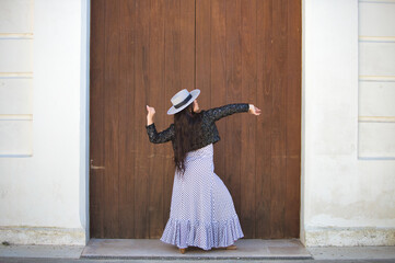 Naklejka premium Latin woman, young and beautiful dancing flamenco in front of a wooden door in a typical street of andalusia. The woman is dressed in a white ruffled dress with black polka dots, a hat and a jacket.