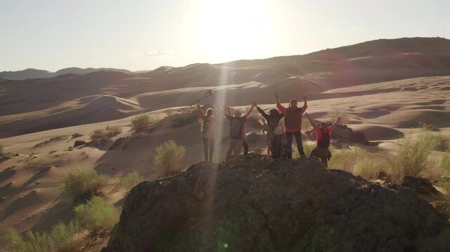A large group of diverse tourists stands on a rock among sand dunes and happily waves their hands, Mongol Els region, Mongolia