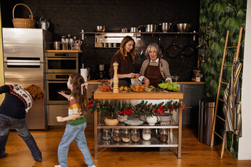 Family, mother and grandmother standing at the table and talking while daughter walks next to them and holds hand towards son, in the kitchen, during Christmas