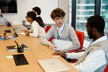 Male intern sitting at a desk and talking to a male employee sitting next to him and holding a pencil, in an office, during an internship