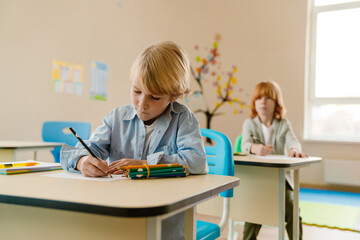 A schoolboy sits at a desk and writes with a pencil while a schoolboy sits behind him and looks...