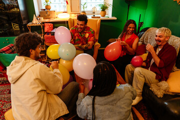 Group of five friends sitting on sofas and chairs and laughing while blowing up balloons, at a party