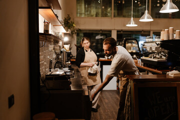 A male employee stands and opens the dishwasher while the female owner stands next to him looks at...