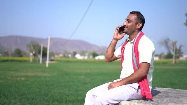 Young happy rural Indian farmer talking on smart phone while sitting at agriculture field. Prosperity and people of india.