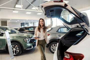 Portrait of a happy female car dealer standing at car salon with tablet in hands and smiling at the...