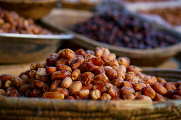 A close-up of fresh dates stacked in a woven basket at an outdoor market, with more dried fruits in the background.