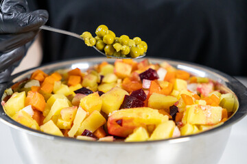 adding green peas to a homemade vinaigrette salad with beets, potatoes, carrots, and pickles in a metal bowl, chef in black gloves