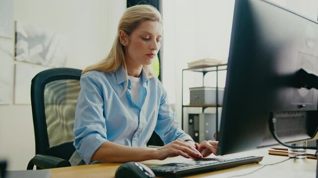 Focused Caucasian woman wearing shirt sitting at desk and typing on keyboard. Concentrating while working on project or responding to corporate emails. Modern workspace with large monitor.