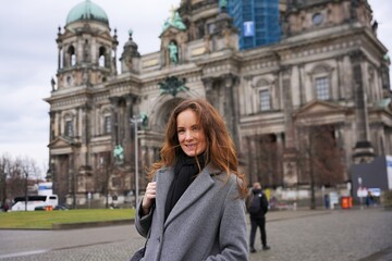 Obraz premium A happy woman in a gray coat and black scarf poses in front of Berlin Cathedral, enjoying the historic architecture and city atmosphere. 