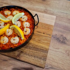 High-angle close-up view of a paella dish on a wooden cutting board. The paella, in a dark-colored metal pan, contains a mixture of rice, shrimp, and vegetables with slices of lemon on top.