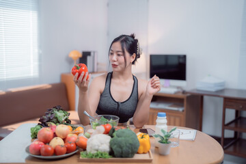 Beautiful asian sportswoman sitting at the table in her living room, choosing a red bell pepper among many other vegetables and fruits, enjoying a healthy lifestyle