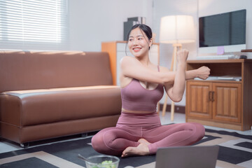 Asian sportswoman sitting in lotus pose on yoga mat and stretching arms while watching online...