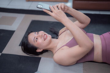 Obraz premium Asian sportswoman in sportswear using mobile phone while relaxing on yoga mat after exercising at home, enjoying online communication, social media, or browsing internet