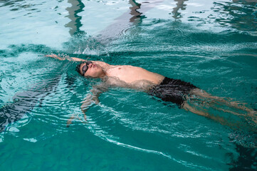 Male Asian swimmer with swim cap and goggles swimming backstroke in pool