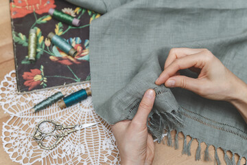 Hands meticulously working on openwork embroidery on grey fabric, surrounded by vintage scissors, thread spools, and a floral book on a lace doily, showcasing a delicate crafting process.