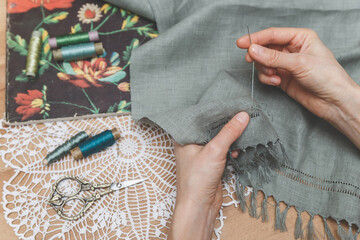 Hands meticulously working on openwork embroidery on grey fabric, surrounded by vintage scissors, thread spools, and a floral book on a lace doily, showcasing a delicate crafting process.