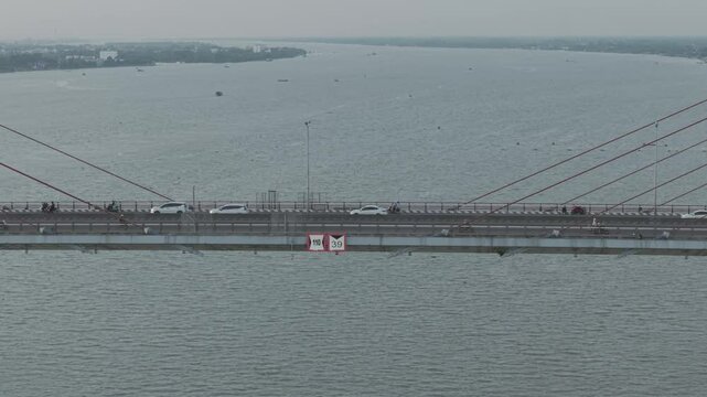A section of Can Tho Bridge spans across the Hau River, Can Tho, Vietnam with cars and motorcycles moving along the roadway, while red cables and speed limit signs are visible in the foreground.