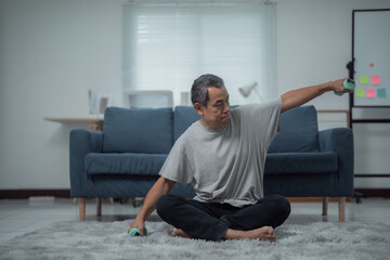 Fototapeta premium Determined senior Asian man lifting light dumbbells while sitting on the floor at home, engaging in exercises for rehabilitation and promoting an active, healthy lifestyle