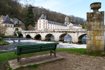 Green bench sits on a lawn near a bridge. It is located near a body of water, crossed by the bridge. Brandonne, France