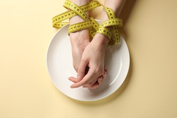 Bulimia. Woman tied with measuring tape and plate on beige background, top view