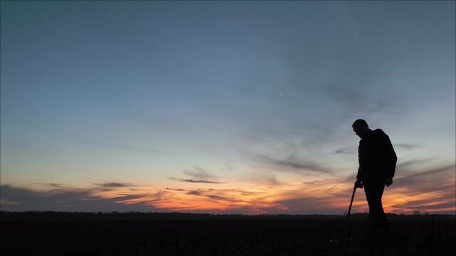 silhouette of a man with a metal detector against the sunset. Silhouette of a man walking along the field with a metal detector against the backdrop of a beautiful sunset.