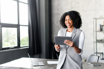 An ebullient African-American professional uses a digital tablet by a sunlit window, her engaging...