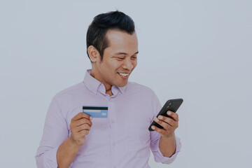 Fototapeta premium Shopping online. Happy asian man holding credit card and mobile phone over white studio background. Empty Space.