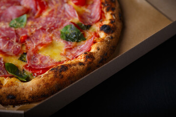 Pizza with cheese, ham, green basil leaves and tomatoes in paper box on black background. Food product advertising banner, copy space
