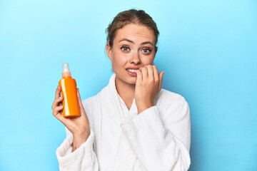 Blonde in bathrobe with sunscreen cream in studio biting fingernails, nervous and very anxious.