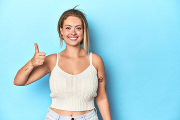 Blonde young woman in white top on blue background smiling and raising thumb up