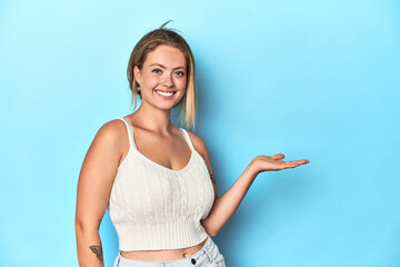 Blonde young woman in white top on blue background showing a copy space on a palm and holding...