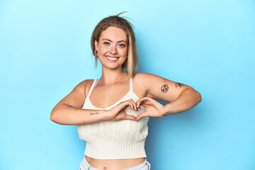 Blonde young woman in white top on blue background smiling and showing a heart shape with hands.