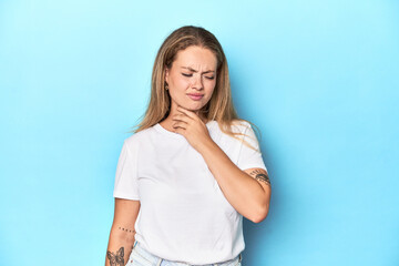 Blonde young woman in white t-shirt on blue background suffers pain in throat due a virus or...