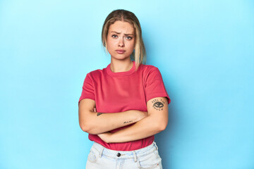 Blonde young woman in red t-shirt on blue background frowning face in displeasure, keeps arms...