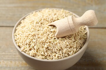 Raw brown rice and scoop in bowl on wooden table, closeup