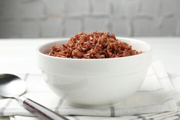 Tasty brown rice in bowl and spoon on white table, closeup